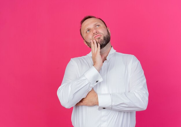 Uomo Barbuto che indossa una camicia bianca che osserva in su con espressione pensierosa sulla faccia pensando in piedi oltre il muro rosa