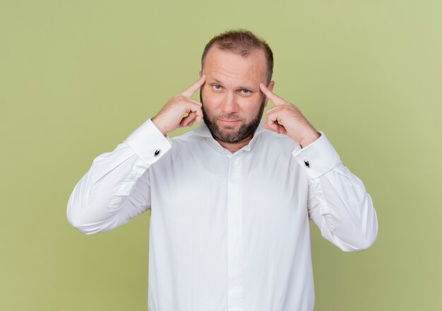 Uomo barbuto che indossa una camicia bianca che punta con il dito indice alle tempie concentrato su un compito difficile in piedi sopra la parete leggera