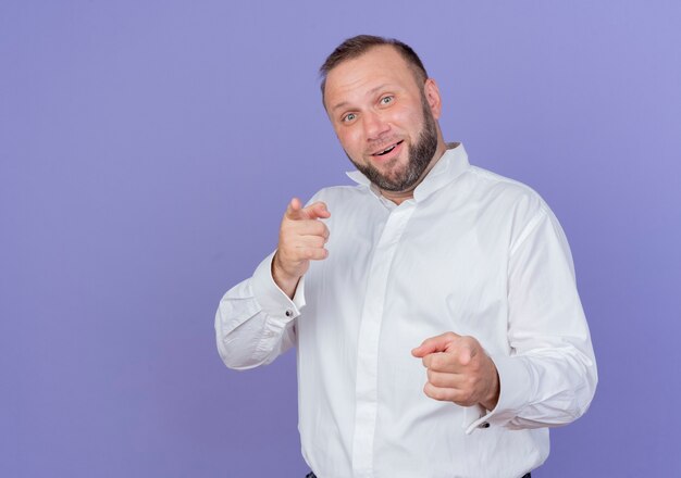 Uomo barbuto che indossa una camicia bianca che punta con le dita indice sorridendo allegramente in piedi sopra la parete blu