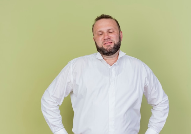 Uomo barbuto che indossa una camicia bianca che sembra scontento con le braccia all'anca in piedi sopra la parete leggera