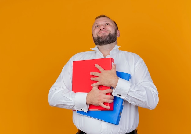 Uomo barbuto che indossa una camicia bianca che tiene le cartelle lookign con la faccia felice in piedi sopra la parete arancione