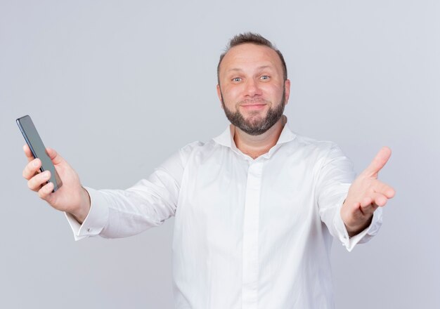 Uomo barbuto che indossa una camicia bianca che tiene smartphone che fa gesto di benvenuto con la mano sorridente in piedi sopra il muro bianco