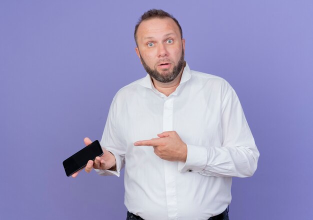Uomo barbuto che indossa una camicia bianca che tiene smartphone che punta con il dito indice contro di essere sorpreso e confuso in piedi sopra la parete blu