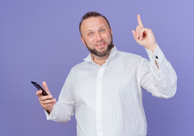 Uomo Barbuto che indossa una camicia bianca che tiene smartphone che sembra felice e sorpreso che mostra il dito indice avente una nuova idea in piedi sopra la parete blu