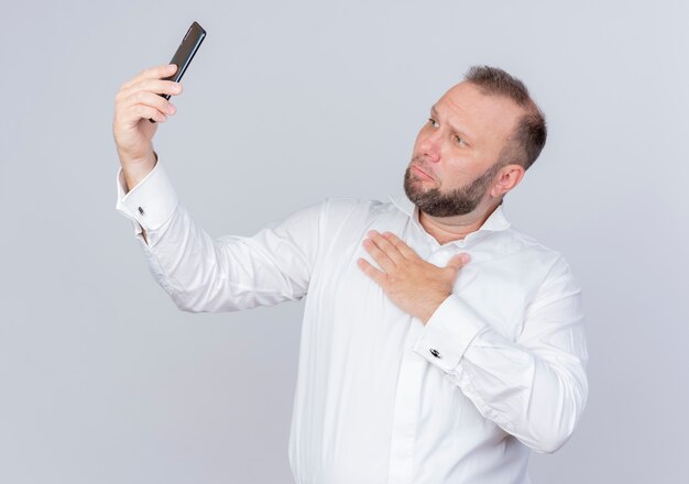 Uomo Barbuto che indossa una camicia bianca che tiene smartphone con videochiamata sentendosi grato tenendo la mano sul petto in piedi sopra il muro bianco