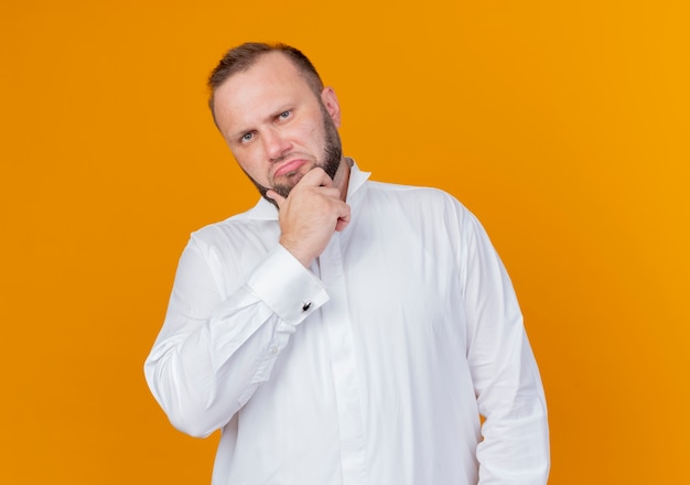 Uomo Barbuto che indossa una camicia bianca con la mano sul mento pensando in piedi sopra la parete arancione