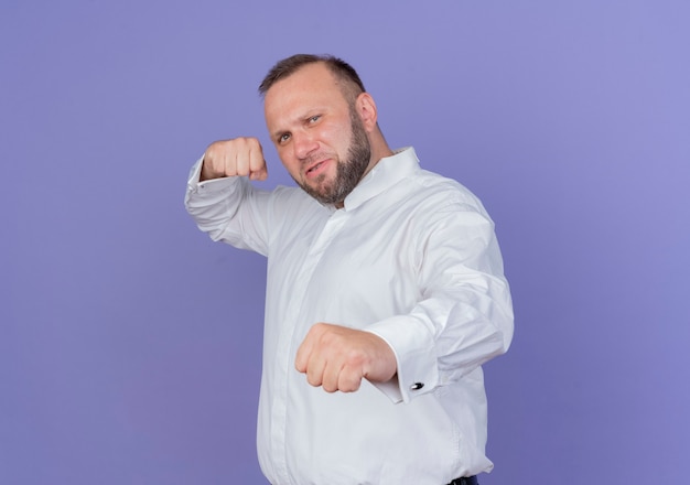 Uomo barbuto che indossa una camicia bianca guardando con faccia seria in posa con il pugno chiuso in piedi sopra la parete blu