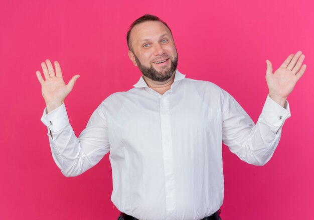Uomo Barbuto che indossa una camicia bianca guardando confuso sorridente alzando le mani in piedi sopra il muro rosa