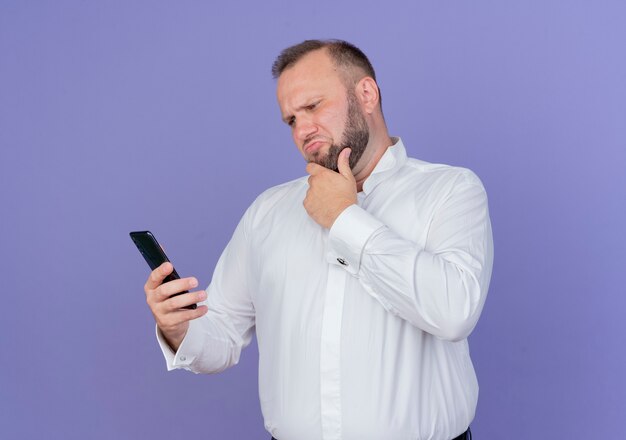 Uomo barbuto che indossa una camicia bianca guardando il suo schermo dello smartphone essendo perplesso in piedi sopra la parete blu