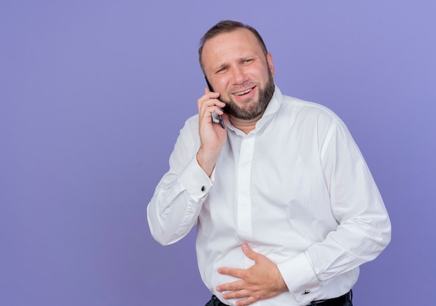 Uomo barbuto che indossa una camicia bianca luaghing mentre parla al telefono cellulare in piedi sopra la parete blu