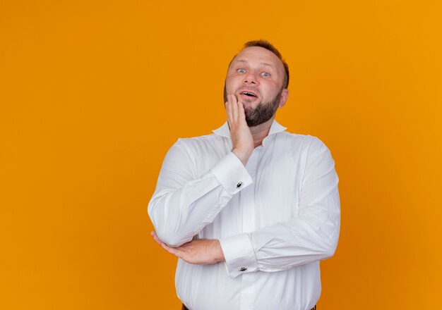 Uomo barbuto che indossa una camicia bianca perplesso in piedi sopra il muro arancione