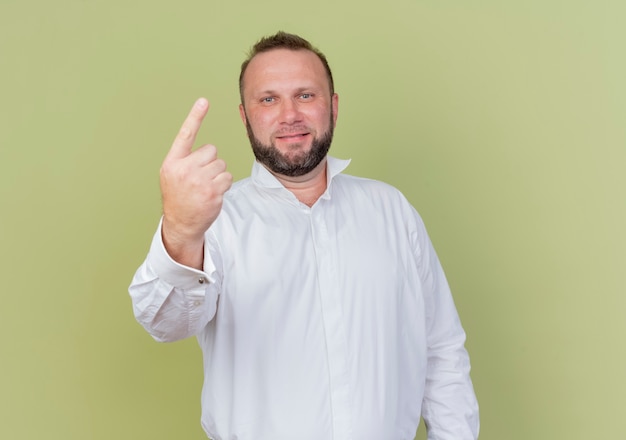 Uomo barbuto che indossa una camicia bianca sorridente che mostra il dito indice o il numero uno in piedi sopra la parete leggera