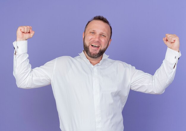 Uomo barbuto che indossa una camicia bianca stringendo i pugni felici ed eccitati in piedi sopra la parete blu