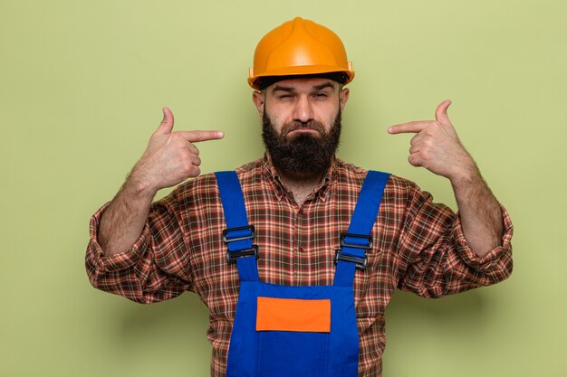 Uomo barbuto costruttore in uniforme da costruzione e casco di sicurezza che guarda con espressione fiduciosa che punta a se stesso