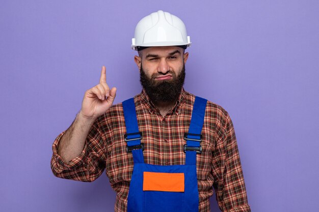 Uomo barbuto costruttore in uniforme da costruzione e casco di sicurezza che guarda la telecamera con espressione sicura che mostra il dito indice in piedi su sfondo viola purple