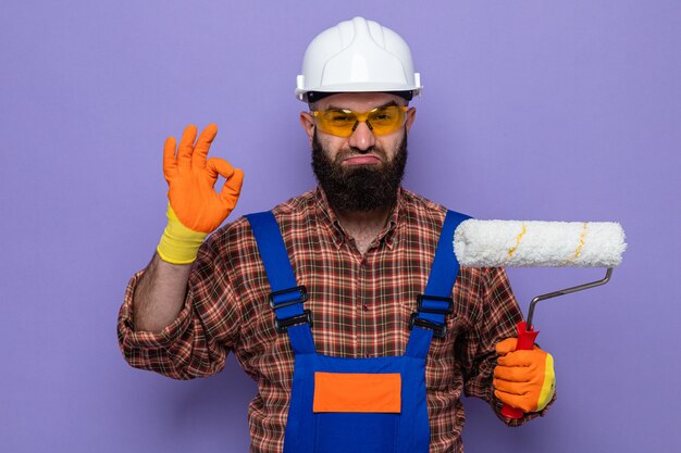 Uomo barbuto costruttore in uniforme da costruzione e casco di sicurezza che indossa guanti di gomma che tengono rullo di vernice guardando la telecamera felice e fiducioso che mostra segno ok in piedi su sfondo viola