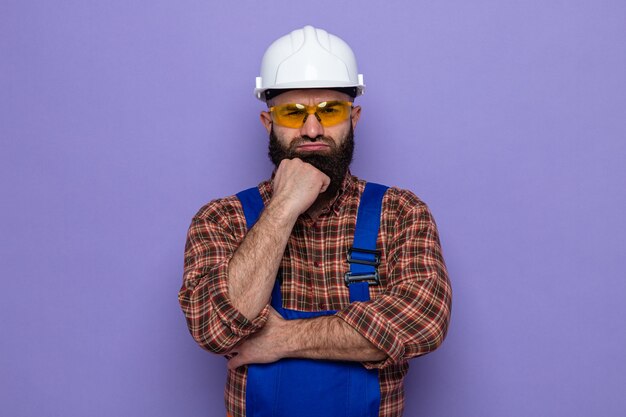 Uomo barbuto costruttore in uniforme da costruzione e casco di sicurezza che indossa occhiali di sicurezza gialli che guarda con la mano sul mento con la faccia arrabbiata