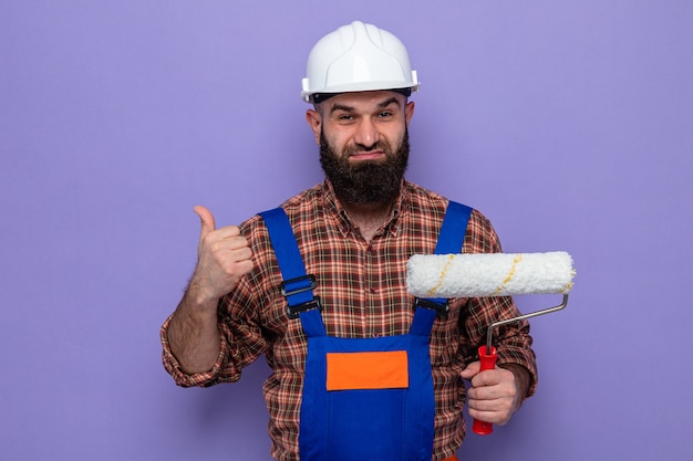 Uomo barbuto costruttore in uniforme da costruzione e casco di sicurezza che tiene il rullo di vernice che sembra sorridente fiducioso che mostra i pollici in su