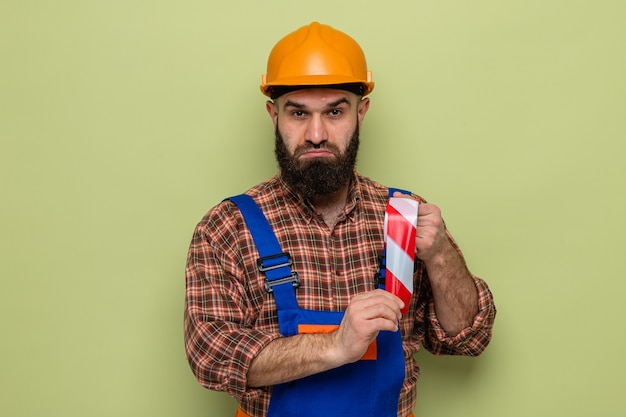 Uomo barbuto costruttore in uniforme da costruzione e casco di sicurezza che tiene nastro adesivo guardando la telecamera confusa in piedi su sfondo verde