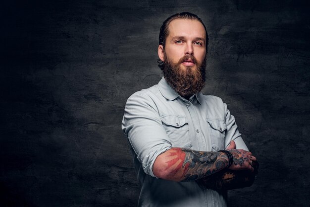 Uomo barbuto curato con tatuaggi sta posando in uno studio fotografico scuro.