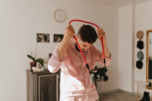 Uomo barbuto dai capelli scuri in camicia leggera che tiene la fotocamera. Ritratto di ragazzo in soggiorno luminoso su sfondo di foto di matrimonio.