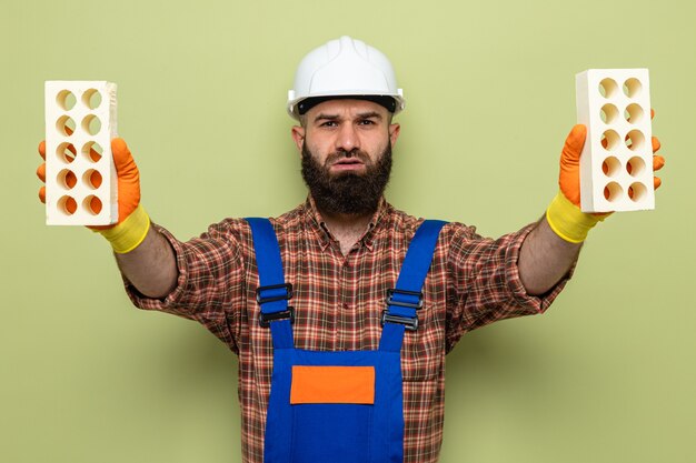 Uomo barbuto del costruttore in uniforme da costruzione e casco di sicurezza che indossa guanti di gomma che tengono i mattoni che guarda l'obbiettivo con la faccia seria che sta sopra fondo verde