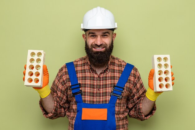 Uomo barbuto del costruttore in uniforme da costruzione e casco di sicurezza che indossa guanti di gomma che tengono i mattoni guardando la telecamera sorridendo allegramente felice e positivo in piedi su sfondo verde