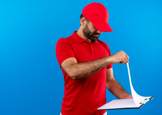 Uomo barbuto di consegna in uniforme rossa e berretto che tiene appunti con pagine bianche guardandoli con faccia seria in piedi sopra il muro blu