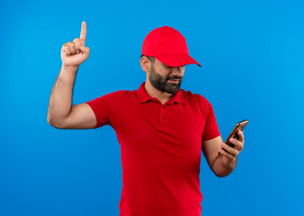 Uomo barbuto di consegna in uniforme rossa e berretto guardando lo schermo del suo cellulare che mostra il dito indice guardando fiducioso in piedi sopra la parete blu