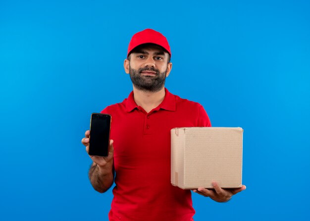 Uomo barbuto di consegna in uniforme rossa e cappuccio che tiene aperto la scatola della pizza che mostra smartphone con espressione sicura in piedi sopra la parete blu