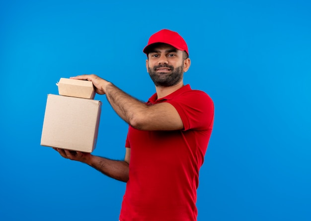 Uomo barbuto di consegna in uniforme rossa e cappuccio che tiene i pacchetti della scatola con un sorriso fiducioso in piedi sopra la parete blu