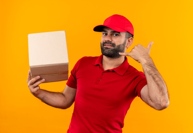 Uomo barbuto di consegna in uniforme rossa e cappuccio che tiene il pacchetto della scatola che mi chiama gesto sorridente in piedi sopra la parete arancione