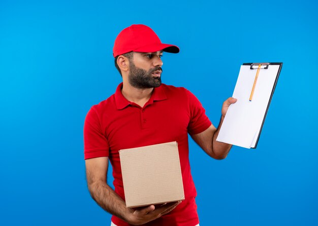 Uomo barbuto di consegna in uniforme rossa e cappuccio che tiene il pacchetto della scatola che mostra appunti con pagine vuote che sembrano confuse in piedi sopra la parete blu