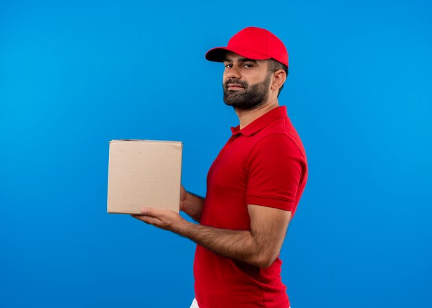 Uomo barbuto di consegna in uniforme rossa e cappuccio che tiene il pacchetto della scatola che sembra fiducioso in piedi lateralmente sopra la parete blu