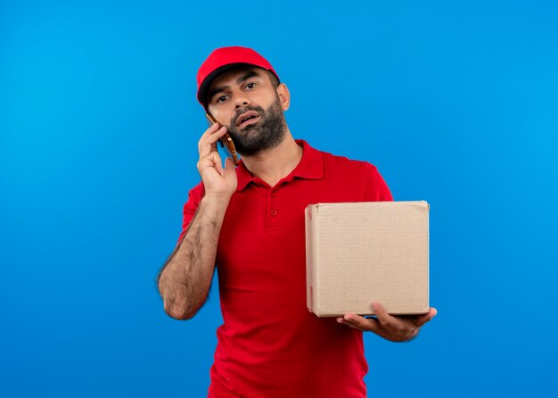 Uomo barbuto di consegna in uniforme rossa e cappuccio che tiene il pacchetto della scatola parlando al telefono cellulare con la faccia seria in piedi sopra la parete blu