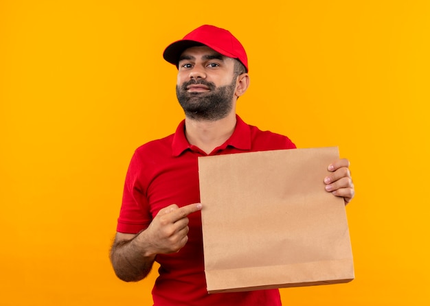 Uomo barbuto di consegna in uniforme rossa e cappuccio che tiene il pacchetto di carta puntato con il dito verso di esso con un sorriso sicuro in piedi sopra la parete arancione