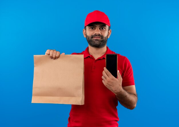 Uomo barbuto di consegna in uniforme rossa e cappuccio che tiene il pacchetto di carta sorridente che mostra smartphone in piedi sopra la parete blu