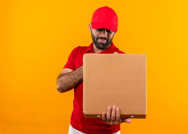 Uomo barbuto di consegna in uniforme rossa e cappuccio che tiene la scatola della pizza aperta guardandolo con una faccia buffa in piedi sopra la parete arancione