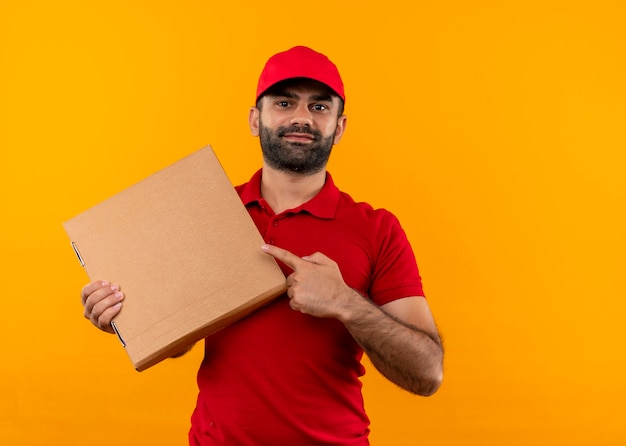 Uomo barbuto di consegna in uniforme rossa e cappuccio che tiene la scatola della pizza che punta con il dito ad essa sorridente fiducioso in piedi sopra la parete arancione