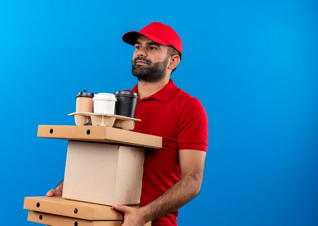 Uomo barbuto di consegna in uniforme rossa e cappuccio che tiene le scatole di cartone che osserva da parte con il sorriso sul viso in piedi sopra la parete blu