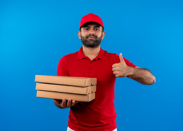 Uomo barbuto di consegna in uniforme rossa e cappuccio che tiene pila di scatole per pizza che mostra i pollici in su sorridente fiducioso in piedi sopra la parete blu