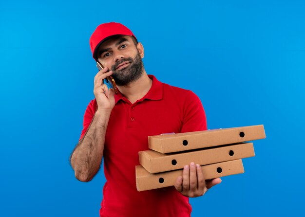 Uomo barbuto di consegna in uniforme rossa e cappuccio che tiene pila di scatole per pizza sorridente mentre parla al telefono cellulare in piedi sopra la parete blu