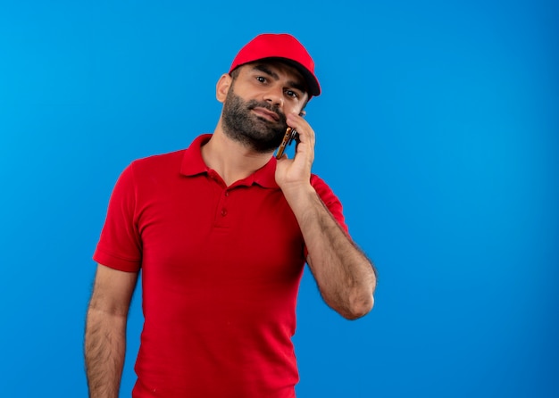 Uomo barbuto di consegna in uniforme rossa e cappuccio parlando al telefono cellulare cercando fiducioso in piedi oltre la parete blu