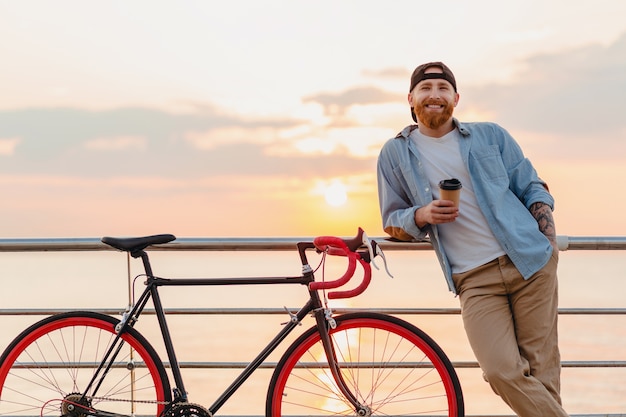 Uomo barbuto di stile hipster felice sorridente bello che indossa camicia di jeans e berretto con la bicicletta all'alba di mattina in riva al mare, bere caffè, viaggiatore sano stile di vita attivo