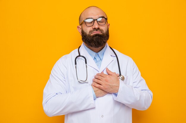 Uomo barbuto dottore in camice bianco con stetoscopio intorno al collo con gli occhiali sorride con gli occhi chiusi tenendosi per mano sul petto sentendosi grato