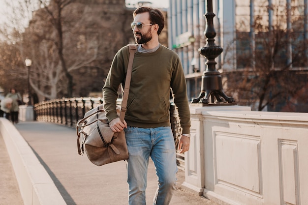 Uomo barbuto elegante bello che cammina nella strada della città con borsa da viaggio in pelle che indossa felpa e occhiali da sole, tendenza di stile urbano, giornata di sole, fiducioso e sorridente