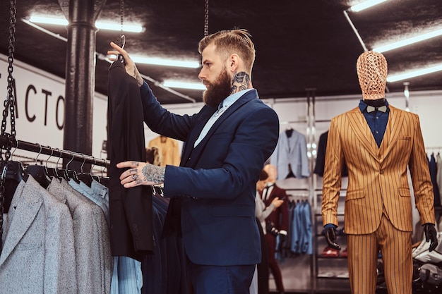 Uomo barbuto elegantemente vestito con tatuaggi su mani e collo sceglie un nuovo abito in un negozio di abbigliamento maschile.