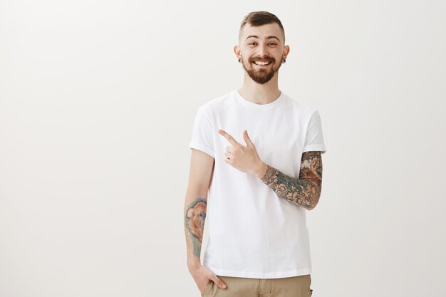 Uomo barbuto felice sorridente con tatuaggi e dito puntato piercing a sinistra