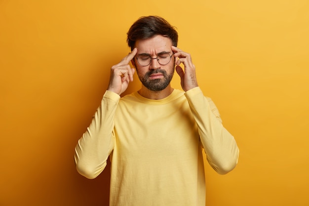 Uomo barbuto frustrato oberato di lavoro massaggia le tempie, soffre di forte emicrania, chiude gli occhi per alleviare il dolore, indossa occhiali da vista e maglione giallo casual, si alza in piedi, cerca di calmarsi