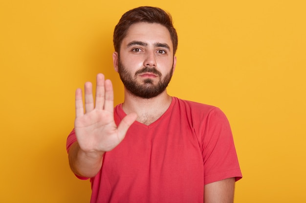 Uomo barbuto giovane che indossa la maglietta casuale rossa che sta con la mano di gesto di avvertimento di arresto e che esamina macchina fotografica con il fronte serio